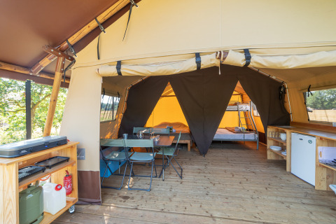 Vista interna di una tenda safari con pavimento in legno, area pranzo, cucina e letti a castello in fondo.