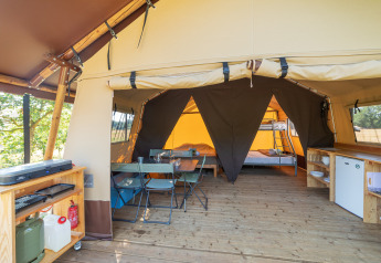 Binnenkant van een safaritent met houten vloer, eethoek, keukentje en stapelbedden achteraan in beeld.