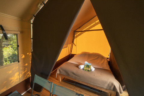 Intérieur d'une tente safari avec un lit double et deux chaises, baigné par la lumière naturelle.