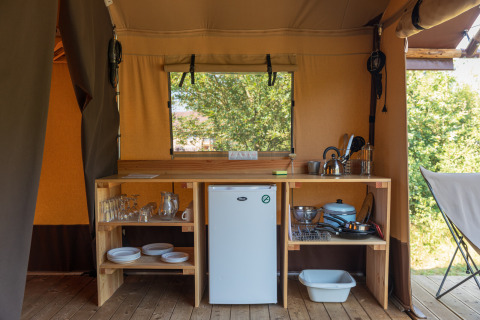 Keukengedeelte in een safaritent met koelkast, houten planken, servies en uitzicht naar buiten.