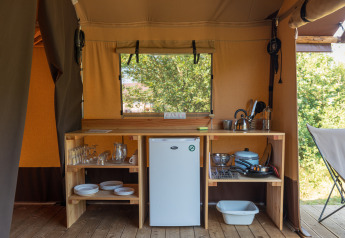 Küchenbereich in einem Safarizelt mit Kühlschrank, Holzmöbeln, Geschirr und Blick ins Grüne.