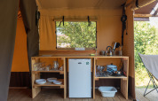 Kitchen area in a safari tent with a fridge, wooden shelves, dishes, utensils, and a window view.