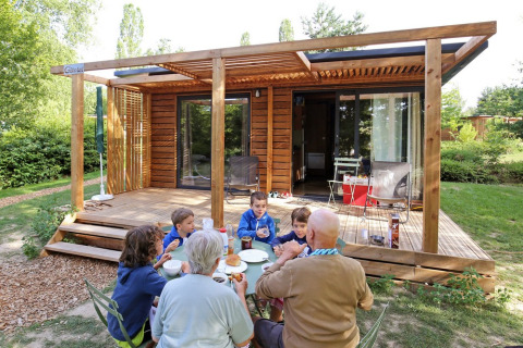 Familia desayunando al aire libre frente a cabaña de madera en Huttopia Etang de Fouché, Borgoña, Francia.