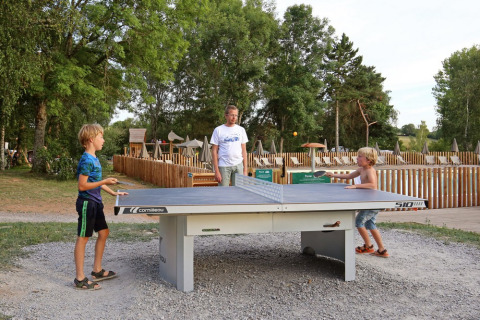 Dos niños juegan al ping-pong al aire libre mientras un adulto observa en Huttopia Etang de Fouché, Francia.