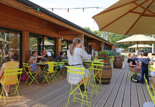 Udendørs caféterrasse på Huttopia Etang de Fouché, afslappede gæster, grønne stole og parasoller.