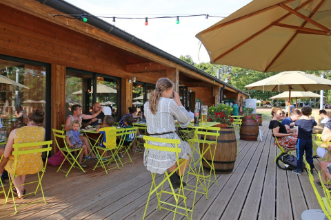 Zonnig terras met gezinnen en groene stoelen bij Huttopia Etang de Fouché vakantiepark in Frankrijk.