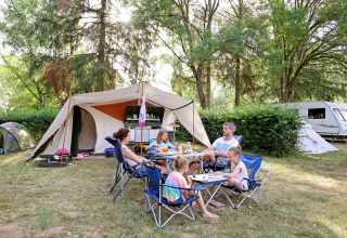 En familie sidder ved et campingbord foran et telt i en grøn skov ved Huttopia Etang de Fouché i Frankrig.