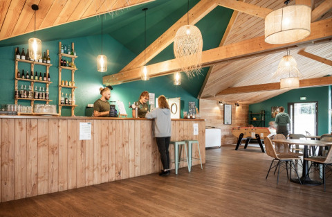 Spacious lounge with wooden beams, bar counter, games area, and cozy lighting at Huttopia Etang de Fouché.