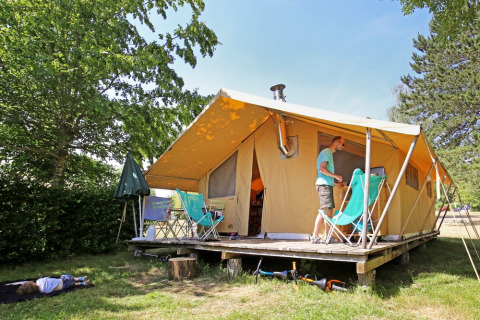 Familie nyder camping udenfor et stort safaritelte med træterrasse i grønne omgivelser ved Huttopia Etang de Fouché.
