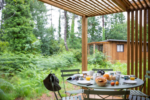 Frühstückstisch auf einer Terrasse beim Chalet Ottawa, Huttopia Etang de Fouché, Frankreich, mit Waldblick.