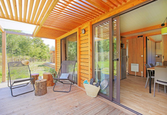 Licht houten chalet met overdekt terras, stoelen en bosrijke omgeving bij Huttopia Etang de Fouché in Frankrijk.