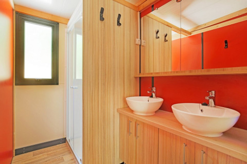 Moderne badkamer met dubbele wastafel, houten kasten en rode accenten in Chalet Ottawa, Huttopia Etang de Fouché.
