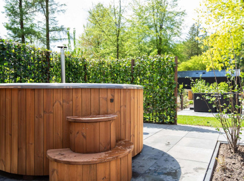 Holz-Whirlpool mit Treppe im Garten des Module Wood Hottub, Bospark Ede, Niederlande, mit grüner Hecke.