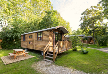 Wooden Gypsy caravan lodge with porch and picnic table, nestled in lush greenery on a sunny day.