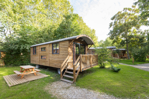 Træbeklædt Gypsy caravan lodge med veranda og picnicbord i grøn, frodig natur om sommeren.