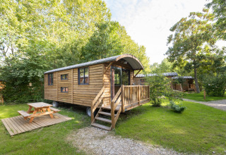Gypsy caravan lodge de madera con porche y mesa de picnic, rodeada de vegetación y naturaleza.