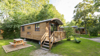 Wooden Gypsy caravan lodge with porch and picnic table, nestled in lush greenery on a sunny day.