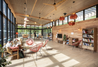 Bright lounge with wooden furniture, large windows, and people relaxing at Camping de Paris, Île de France.