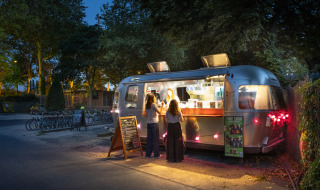 Caravana de comida ubicada en Camping de Paris, Île de France, Francia, con clientes durante la noche.