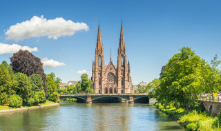 Vista panorámica de una iglesia gótica junto al río en Estrasburgo, Francia, rodeada de árboles verdes.