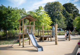 Speelplein met glijbaan en klimconstructie op Camping de Strasbourg, omgeven door bomen en families.