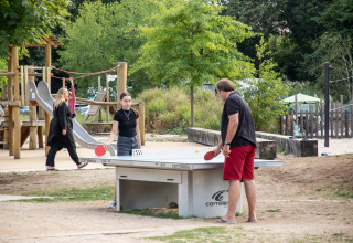 Bezoekers spelen een partijtje tafeltennis buiten op Camping de Strasbourg, met speeltuin op de achtergrond.