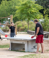 Gäste spielen Tischtennis im Freien im Camping de Strasbourg Ferienpark, mit Spielplatz im Hintergrund.