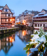 Escena invernal de Estrasburgo, Francia, con casas de entramado de madera y nieve junto a un canal tranquilo.