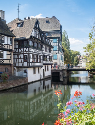 Casas de entramado de madera junto a un canal en Estrasburgo, Francia, con flores y un puente soleado.