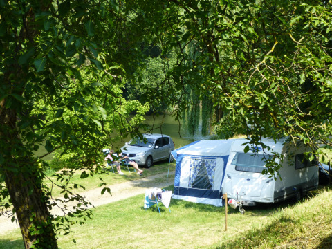 Escena de camping junto a un río con caravana, coche y personas descansando en Camping de l'Ill - Colmar, Francia.