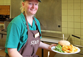Kvinde i personaletøj serverer tallerkener med burger, pommes frites og salat i et køkken på Camping de l'Ill - Colmar.