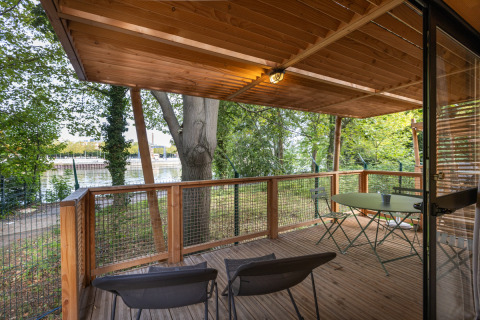 Træterrasse med borde og stole på Chalet Evasion, Camping de Paris, med udsigt til Seinen og træer.