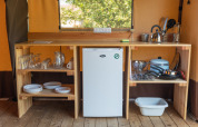 Piccola cucina in una tenda Toile&bois Classic al Camping de Strasbourg con piatti, pentole e frigorifero.