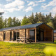 Naturhytte ved Wilsumer Berge i Tyskland, omgivet af træer og græs, med moderne træfacade og udendørs siddeområde.