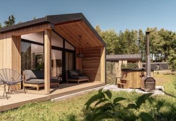 Moderna cabaña de madera con terraza techada, asientos al aire libre y bañera en un entorno natural verde.