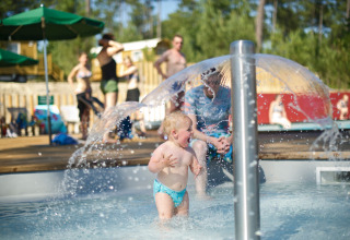 Lille barn leger under vandfontæne i soppebassin med voksne og børn i baggrunden i Huttopia Landes Sud.