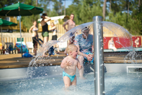 Niño pequeño jugando bajo una fuente de agua en la piscina con familias en Huttopia Landes Sud, Nouvelle-Aquitaine.