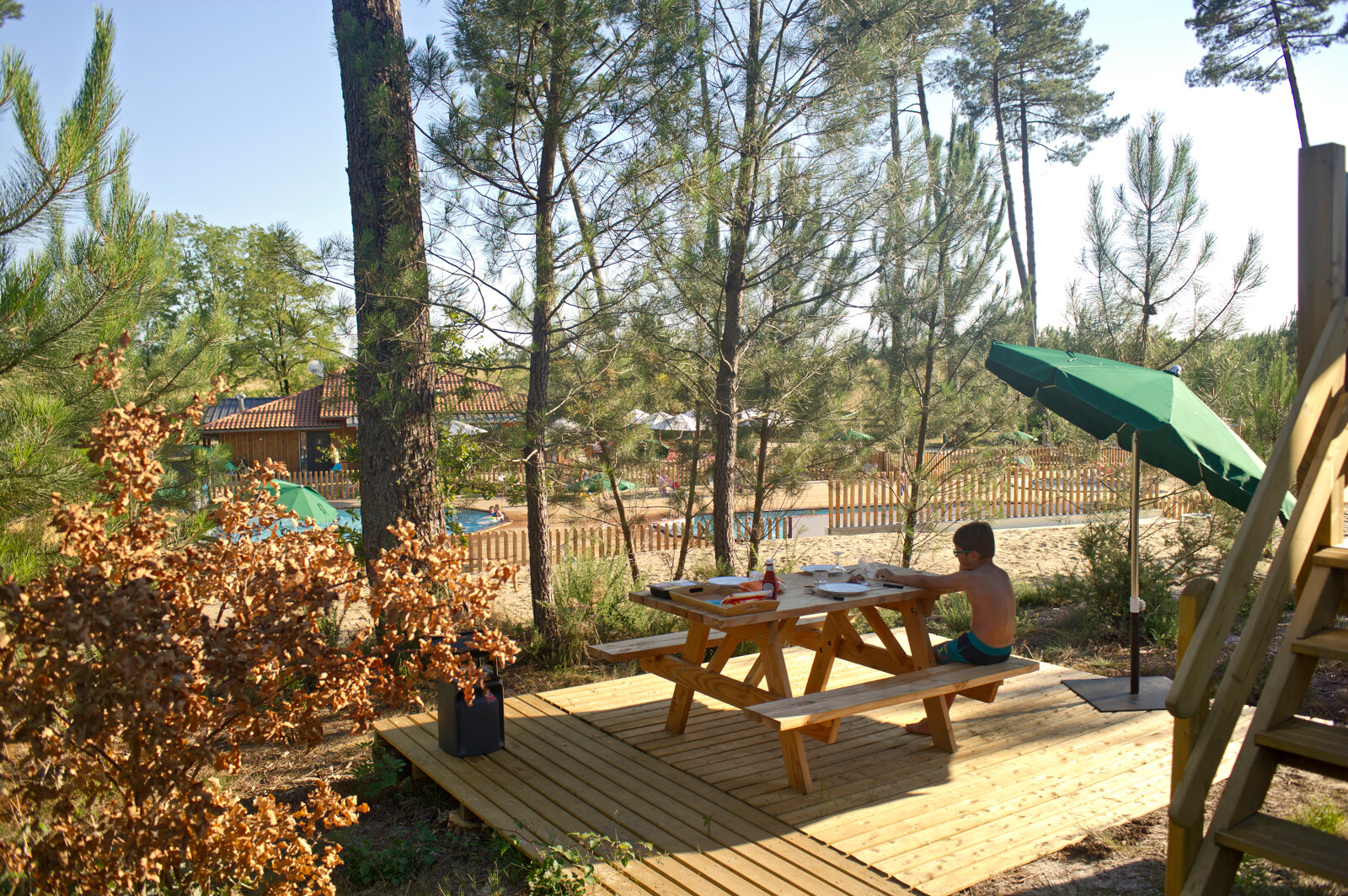 Een jongen zit aan een picknicktafel met parasol bij een hut in het bos bij Huttopia Landes Sud, Frankrijk.