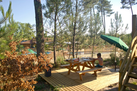 Ein Junge sitzt am Holztisch unter einem Sonnenschirm an einer Waldhütte bei Huttopia Landes Sud, Frankreich.