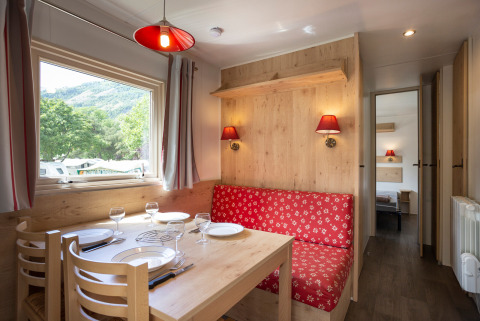 Acogedora cabaña con mesa de comedor, sofá rojo floral, ventana con vista natural y toques de madera.