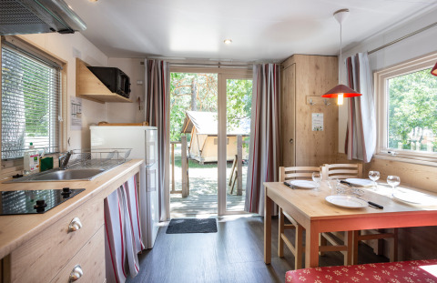 Intérieur de cabane chaleureux avec coin cuisine, table à manger et vue sur la terrasse extérieure boisée.