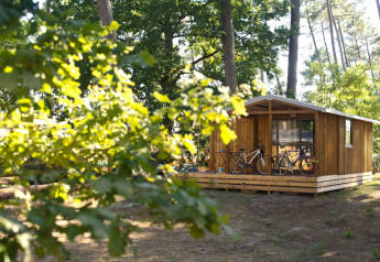 Cabaña Chalet Evasion en Huttopia Landes Sud, Francia, con bicicletas en el porche y rodeada de bosque.