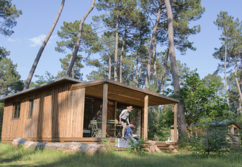 Cabaña de madera Chalet Ottawa en el bosque en Huttopia Landes Sud, Francia, con pareja en la terraza.