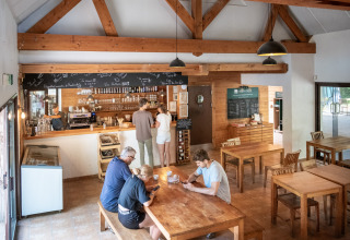 Et lyst caféinteriør i Huttopia Rambouillet med gæster, der sidder ved træborde og bestiller kaffe.