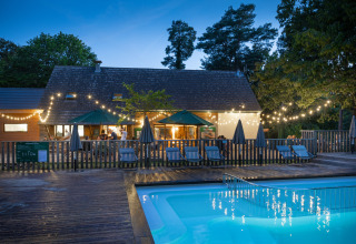 Evening view of Huttopia Rambouillet holiday park with a lit pool, terrace, and string lights in France.