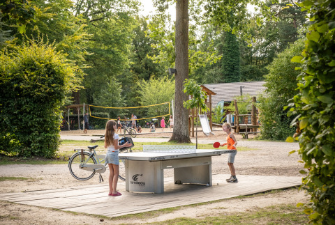 Dos niños juegan al ping-pong al aire libre en Huttopia Rambouillet, con parque infantil y cancha de vóley.