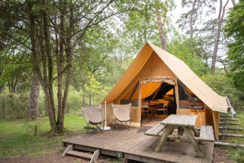 Tenda safari Canadienne con terrazza in legno, due sedie e tavolo da picnic immersi nel verde bosco.