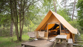 Tenda safari Canadienne con terrazza in legno, due sedie e tavolo da picnic immersi nel verde bosco.