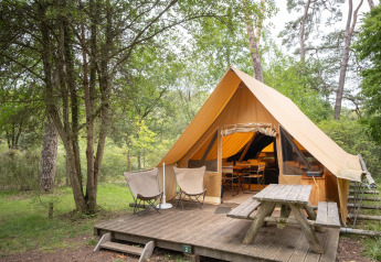 Tenda safari Canadienne con terrazza in legno, due sedie e tavolo da picnic immersi nel verde bosco.