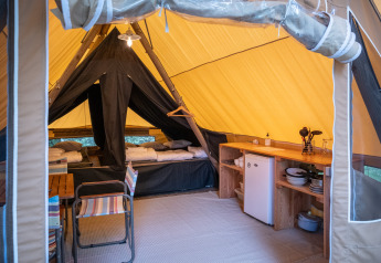 Interno di una tenda safari con letti, zona pranzo e angolo cucina attrezzato con piatti e frigorifero.
